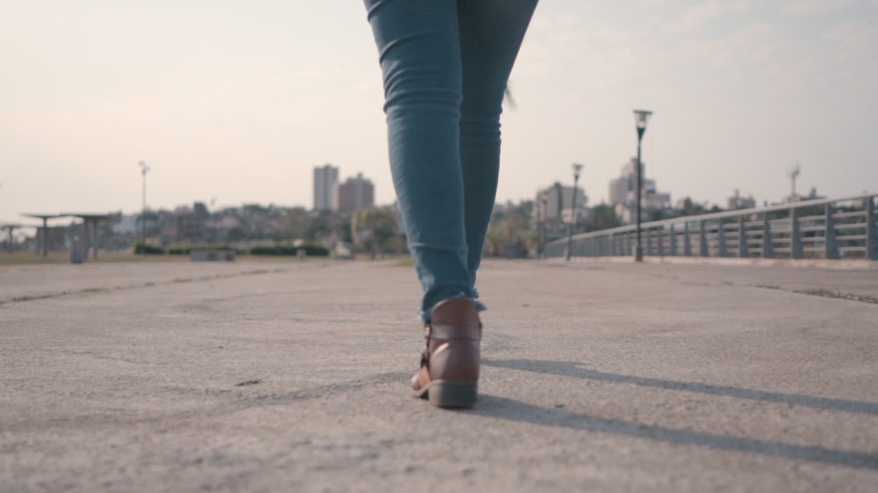 Camera tracking woman's lower body as she walks along an empty street, focused on her stride, in brown boots and jeans