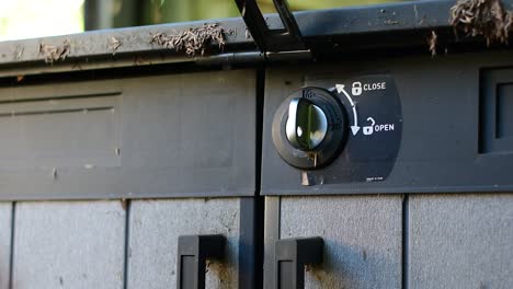 Beautifully-shot-alternate-angle-close-up-of-a-padlock-and-latch-on-a-shed-with-hands-reaching-in-to-use-a-key-to-unlock-and-remove-the-lock-and-unlatch-the-lid-and-door,-with-stylish-bokeh