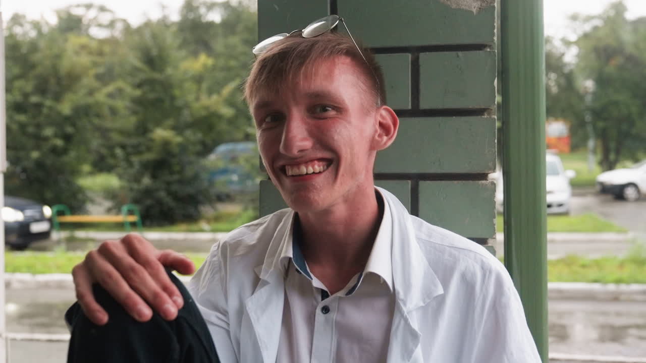 Close view of scientific researcher in white shirt and coat sitting with relaxed posture, smiling warmly outdoors near wall, with glasses on head, reflecting casual human expression and friendly mood