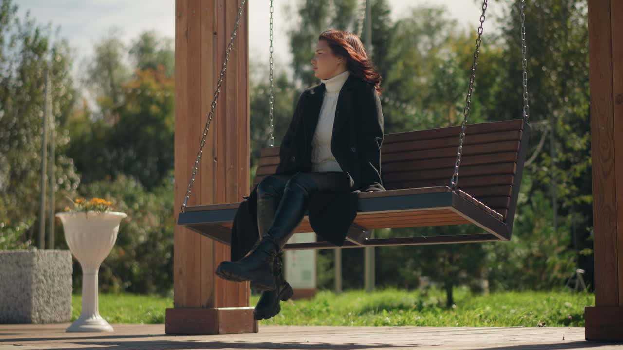 검은 코트와 가죽 부츠를 입은 젊은 여성은 공원에서 나무 스에, 머리카락은 바람에 흔들리고, 은 날 꽃과 초록색으로 둘러싸여 있습니다.