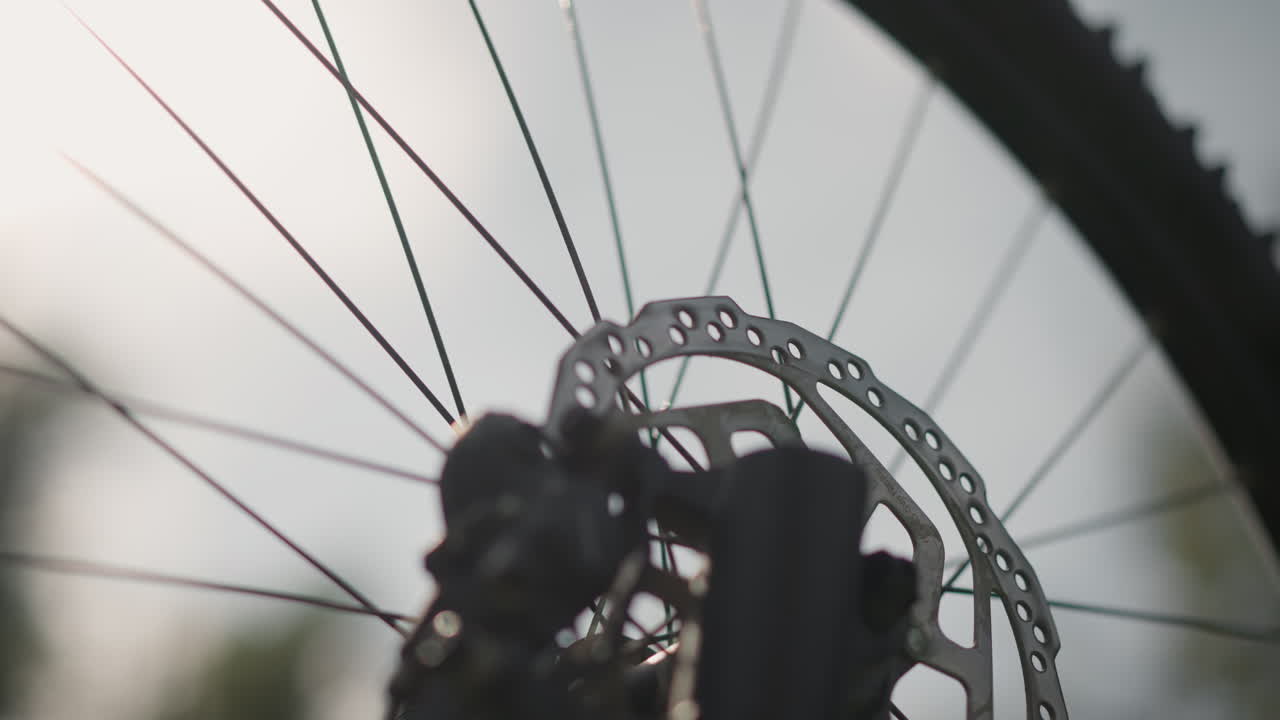 Primo piano del rotore del freno a disco di una bici con raggi e cerchione su cielo pallido, messa a fuoco nitida sulle parti meccaniche, movimento di precisione, ruota da fuoristrada, riflesso della luce solare sul metallo, texture industriale, attrezzatura da ciclismo