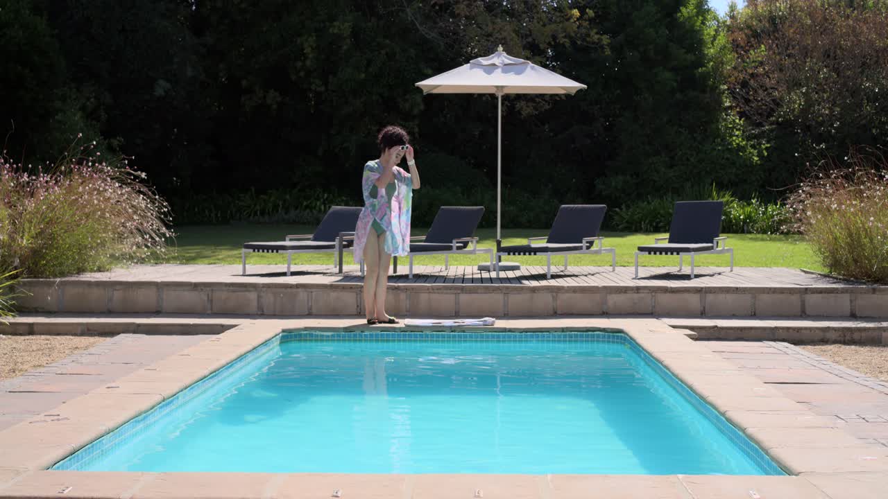 Eine Frau kommt von links herein, trägt ein Handtuch, rollt es am Pool aus, sitzt und taucht ihre Füße ins Wasser, entspannend.