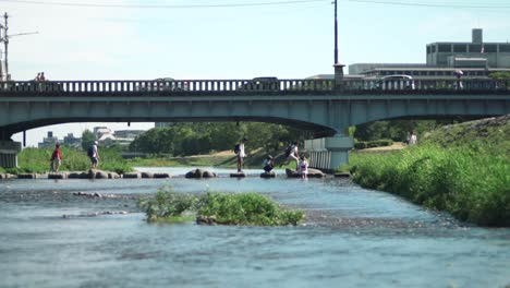 people jumping from rock to rock to cross the kamogawa river with cars travelling on the bridge in the background in kyoto, japan