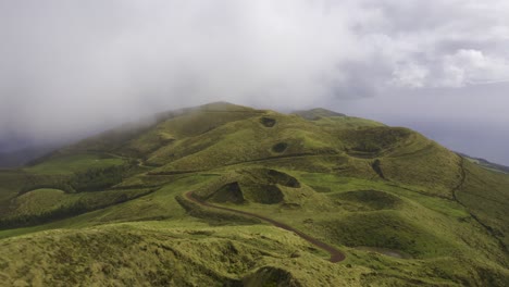 drone-dolly-view-of-vulcanic-mountains-covered-in-lush-green-vegetation-with-dirt-road,-low-clouds-moving-in-São-Jorge-island,-the-Azores,-Portugal