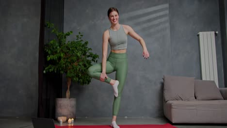 Confident-sporty-brunette-girl-in-a-summer-sports-uniform-with-a-gray-top-and-green-pants-stands-in-a-tree-pose-doing-yoga-on-a-red-sports-mat-near-lit-candles-and-a-houseplant-in-a-modern-apartment