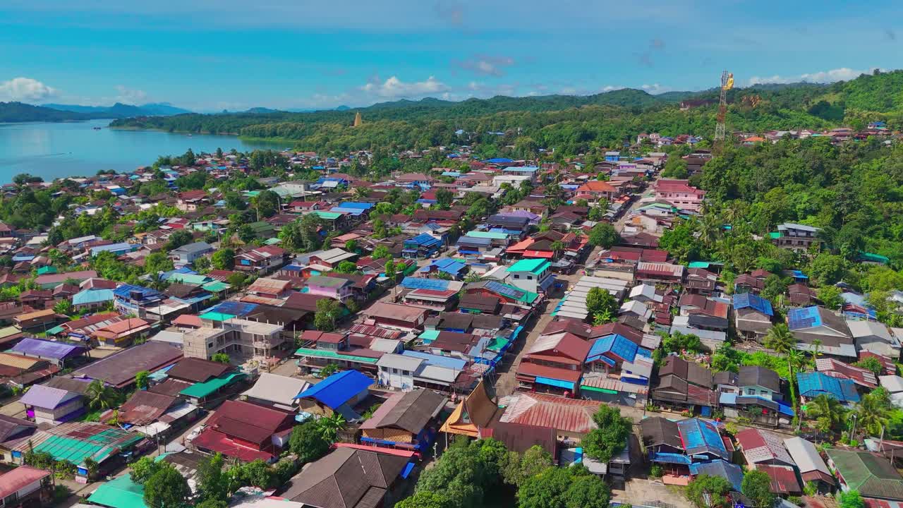 Aerial drone push-in over Mon Village in Sangkhlaburi, Thailand. Colorful rooftops, lush greenery, hillside temples, and a golden Buddha statue in the distance