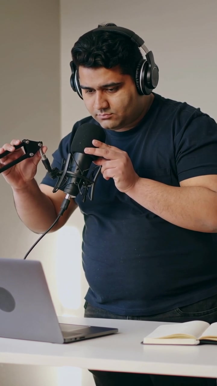 Video verticale: Uomo con maglietta blu scuro che indossa le cuffie al laptop, regolando il microfono in studio per registrare.