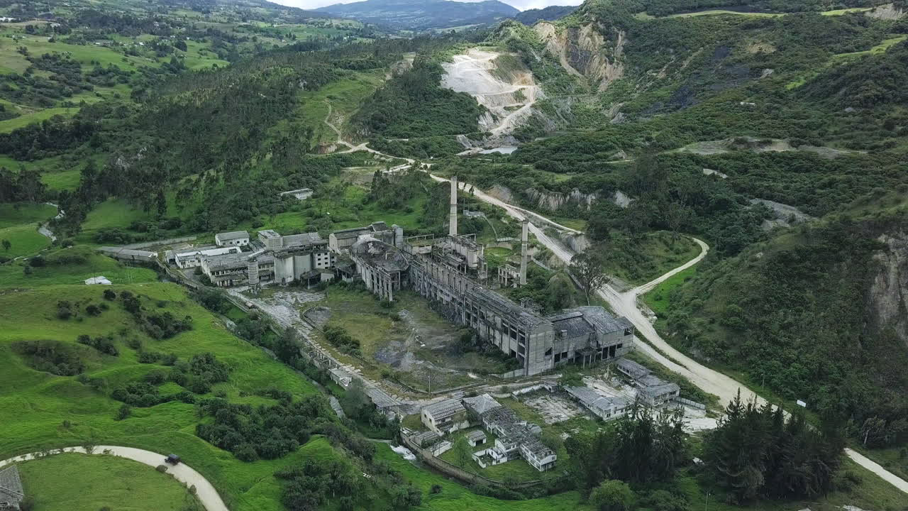 Slow aerial orbit showing the full scale of the abandoned cement plant near La Calera, Colombia. Surrounded by green hills, the crumbling facility contrasts with the natural landscape.