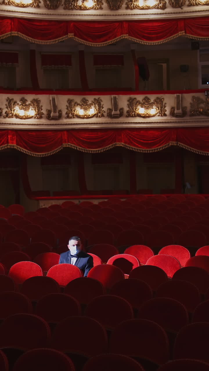 Rich man in mask in empty theater hall. Man spectator is sitting alone in large auditorium and looking to the sides. Consequences of pandemic. Vertical video