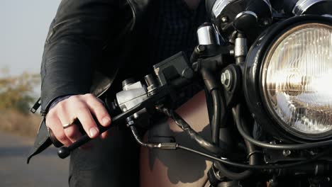 vista frontal de un hombre irreconocible en chaqueta de cuero negro montando una motocicleta en una carretera de campo. gran faro encendido