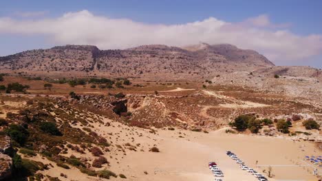 Aufsteigende-Drohnenbilder-Vom-Strand-Von-Rhodos-Und-Den-Vielen-Geparkten-Autos-Mit-Den-Bergen-Im-Hintergrund