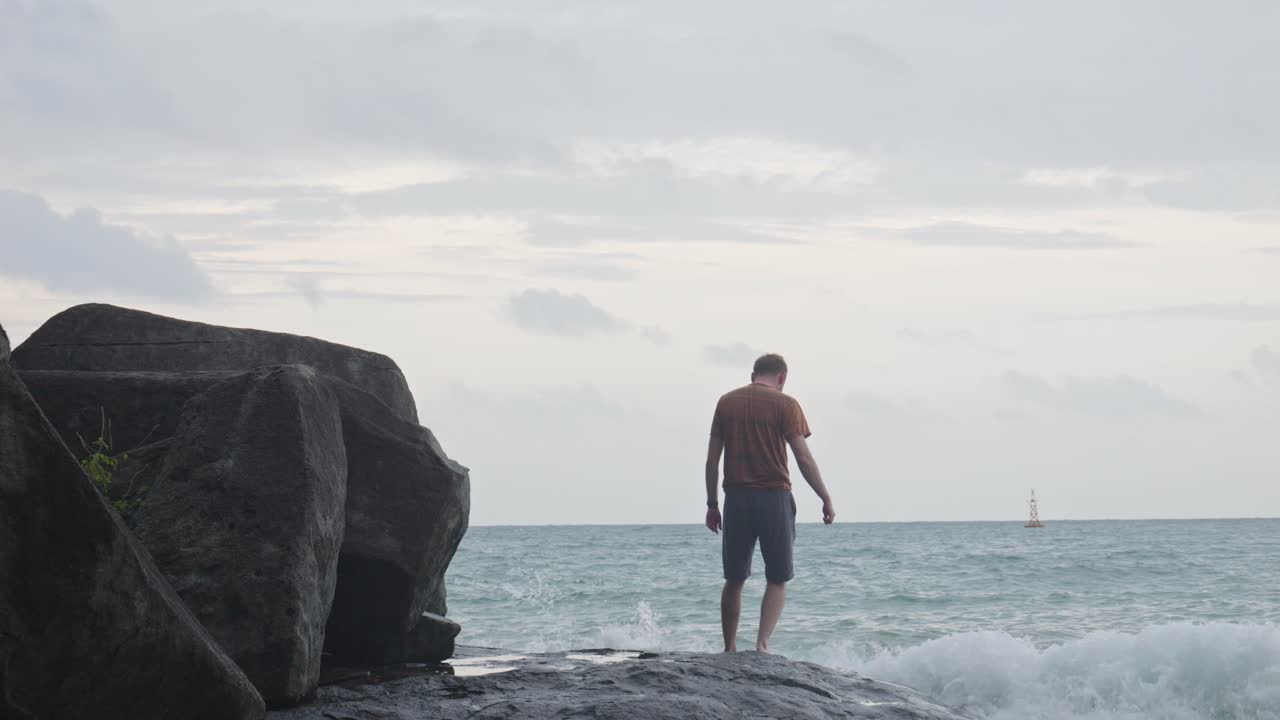 vista detrás de un hombre que va a la playa caminando cuidadosamente sobre una roca húmeda hacia el océano ondulado en la playa de dam trau en vietnam