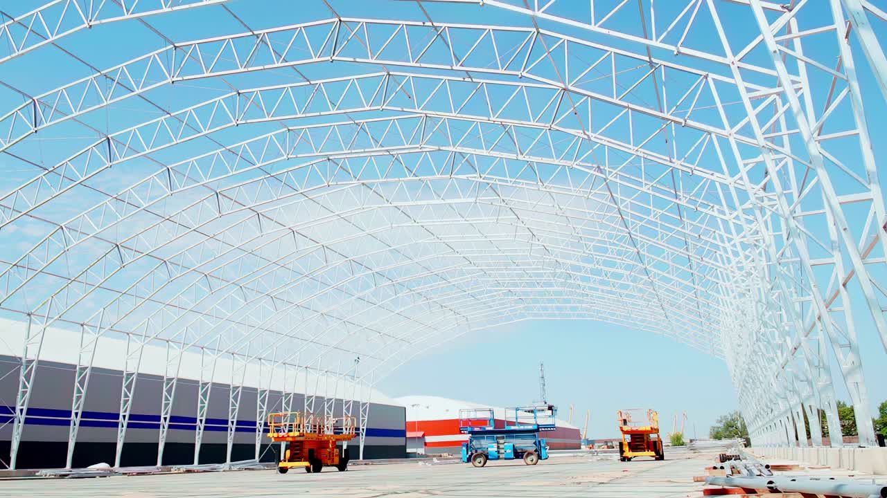 Ampia inquadratura di un grande magazzino con struttura in acciaio in costruzione, con piattaforme di lavoro elevate in un cantiere industriale.