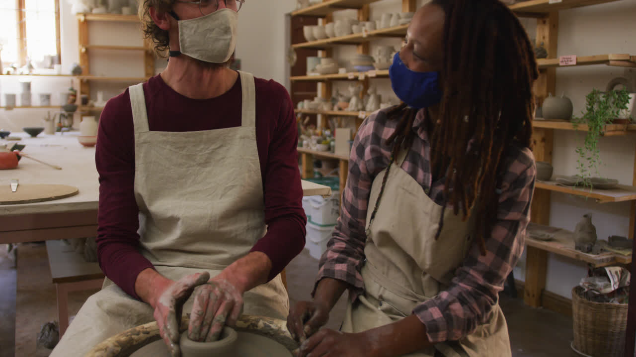 Diverse male and female potters wearing face masks creating pottery on potters wheel at pottery stud