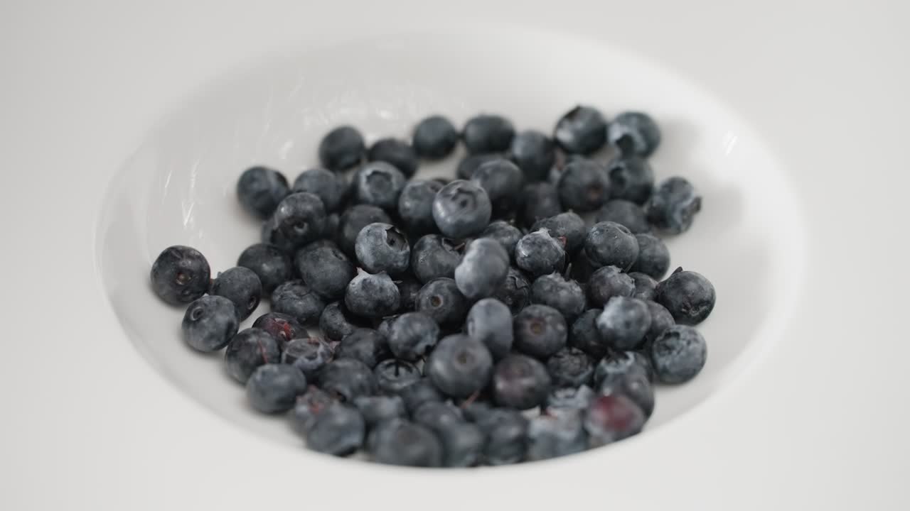 A detailed macro view of plump, organic blueberries dropping and landing in a pristine white dish in super slow-motion, illustrating concepts of wellness, whole foods, and clean eating.