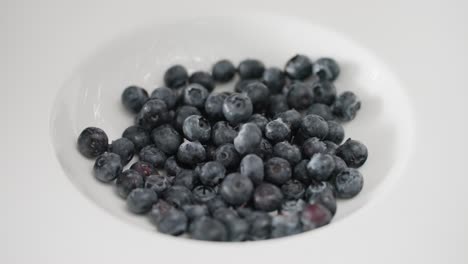 a detailed macro view of plump, organic blueberries dropping and landing in a pristine white dish in super slow-motion, illustrating concepts of wellness, whole foods, and clean eating.