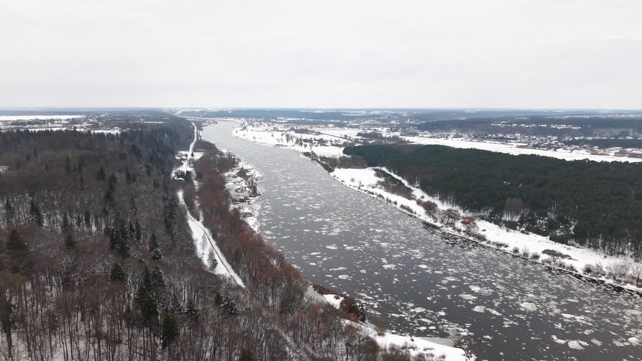 Drone-video, joka kuvaa seesteisen talvimaiseman lumipeitteisen joen ja metsän yllä. Jäätä kelluu vedessä rehevien, ikivihreiden kasvien ympäröimänä luoden rauhallisen ja viehättävän näkymän.