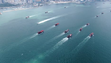 Convoy-of-local-Fishing-boats-causing-in-Hong-Kong-Victoria-bay,-with-city-skyline-in-the-horizon,-Aerial-view