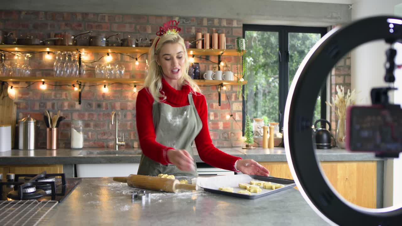 Ring light guiding woman baker adjusting phone, presenting cookie tray for demo on kitchen island