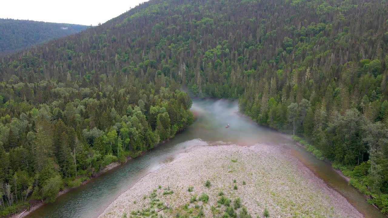 Съемка с дрона запечатлела извилистую реку с галечной косой, окруженную пышным зеленым таежным лесом на склонах гор, с утренним туманом, поднимающимся над водой.