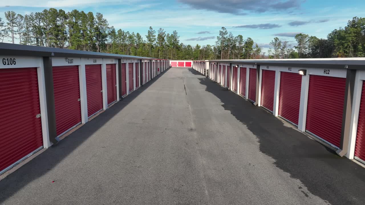Aerial view of an American self-storage facility with long rows of red storage unit doors, clean asphalt lanes and soft daylight. Symmetrical, minimal industrial scene in U.S. Dolly backwards move
