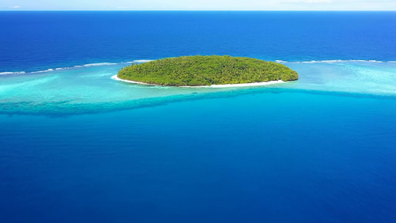 Una vista aerea scivola oltre una piccola isola verde delimitata da sabbia chiara e vivide acque basse turchesi, con formazioni coralline che sfumano in un vasto oceano blu profondo sotto la chiara luce del giorno.