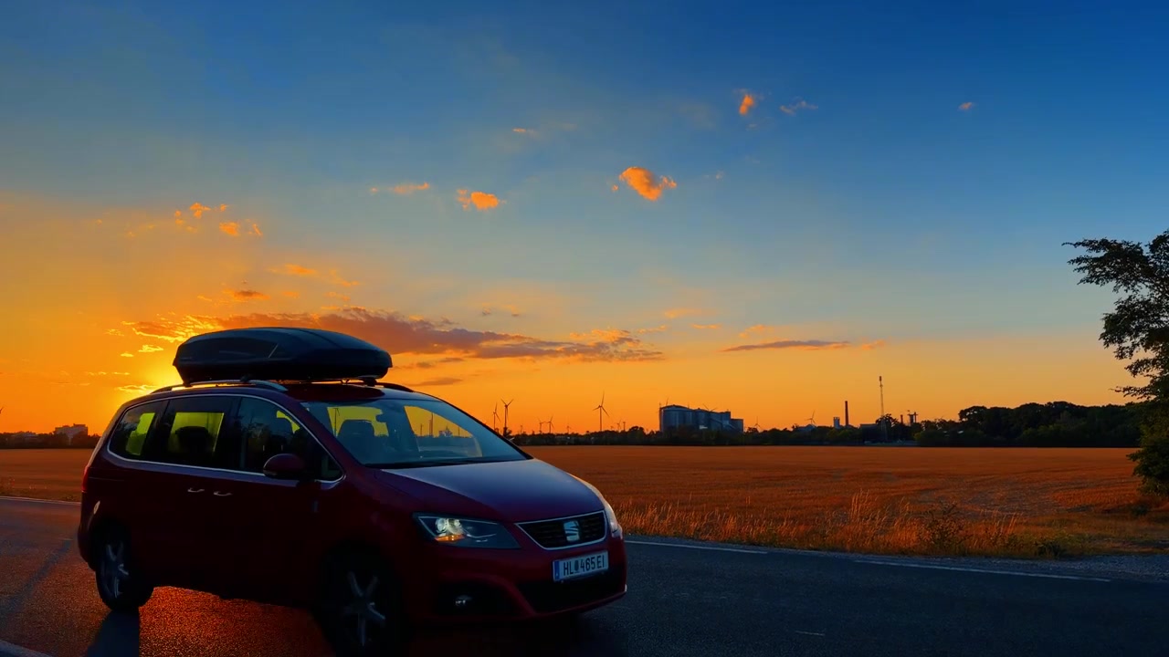 Sole splendente che tramonta su un campo raccolto e l'orizzonte. Sagome di turbine eoliche si ergono lungo l'orizzonte lontano accanto a una strada asfaltata al crepuscolo.