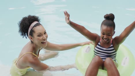 portrait of african american mother and daughter playing in swimming pool in garden, slow motion