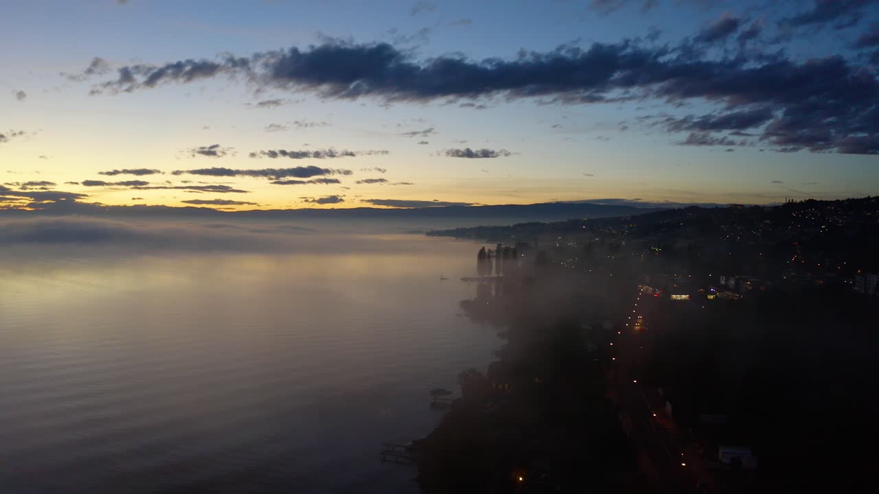 toma aérea que desciende hacia la orilla del lago a través de la niebla, al atardecer con manchas de niebla que se reflejan en el agua lutry, vaud - suiza