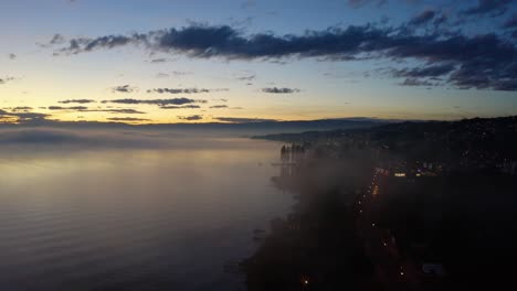 Toma-Aérea-Que-Desciende-Hacia-La-Orilla-Del-Lago-A-Través-De-La-Niebla,-Al-Atardecer-Con-Manchas-De-Niebla-Que-Se-Reflejan-En-El-Agua-Lutry,-Vaud---Suiza