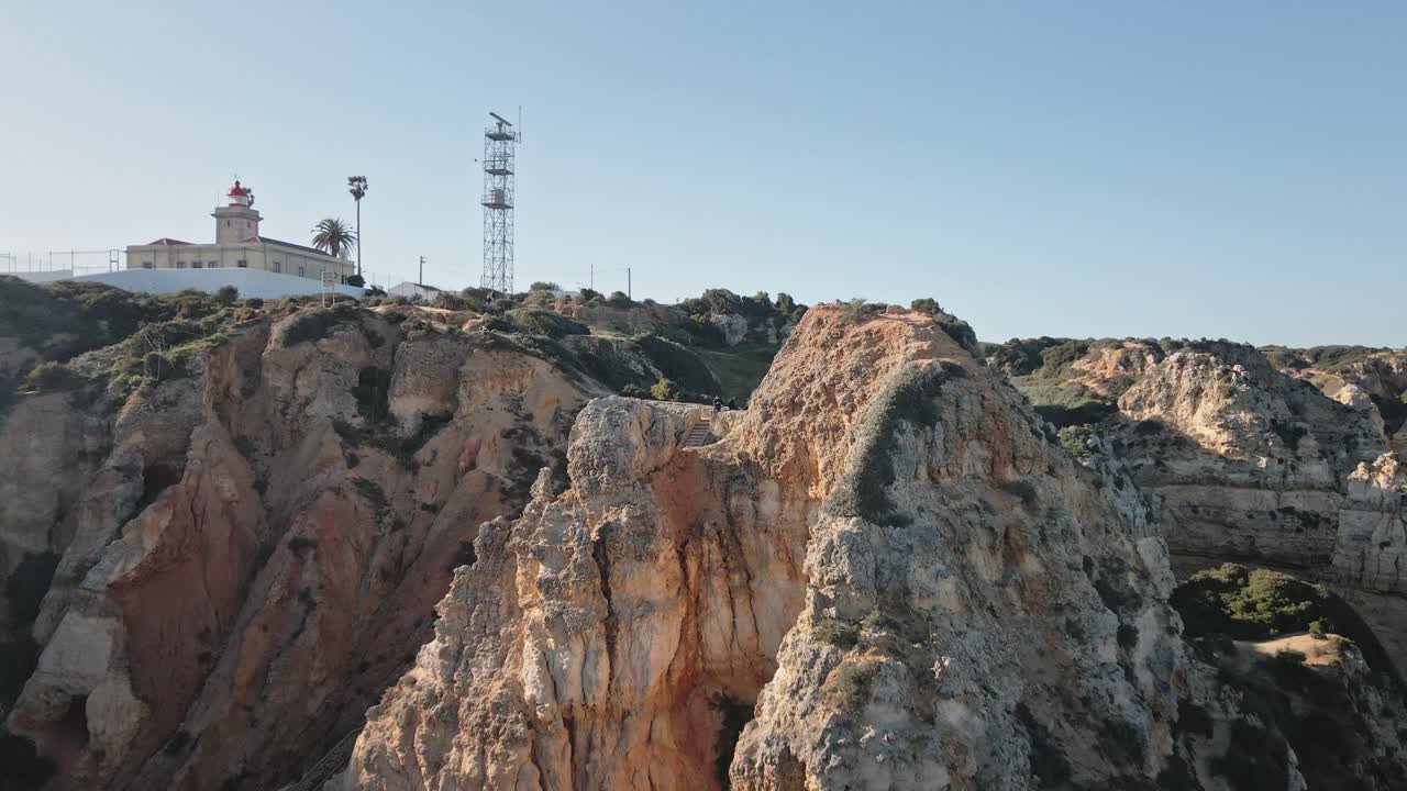 Camera glides back from rugged cliffs, gradually unveiling a dramatic coastal landscape with lighthouse, lookout stairs, and blue sky