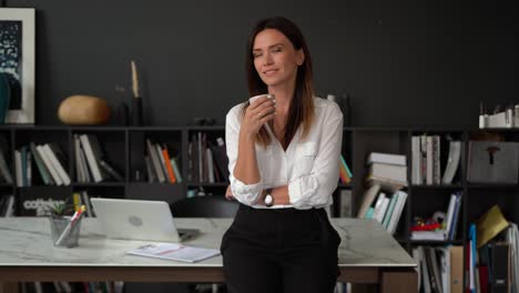 mujer europea joven adulta sonriente y confiada mirando a la cámara de pie en la oficina, posando para un retrato de cerca en el interior