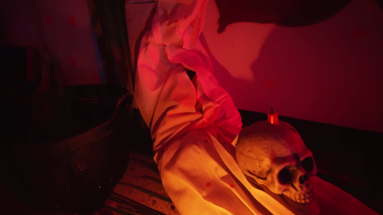 Stanza decorata per una festa di Halloween con un teschio adagiato su un corpo senza vita, candele accese e illuminazione rossa.
