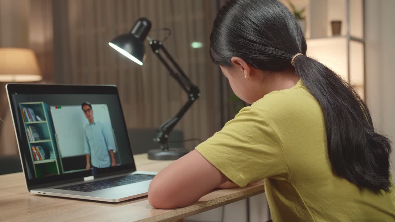 Back View Of Asian Kid Girl Sitting In A Wheelchair While Learning Online With Teacher On Laptop Computer At Home
