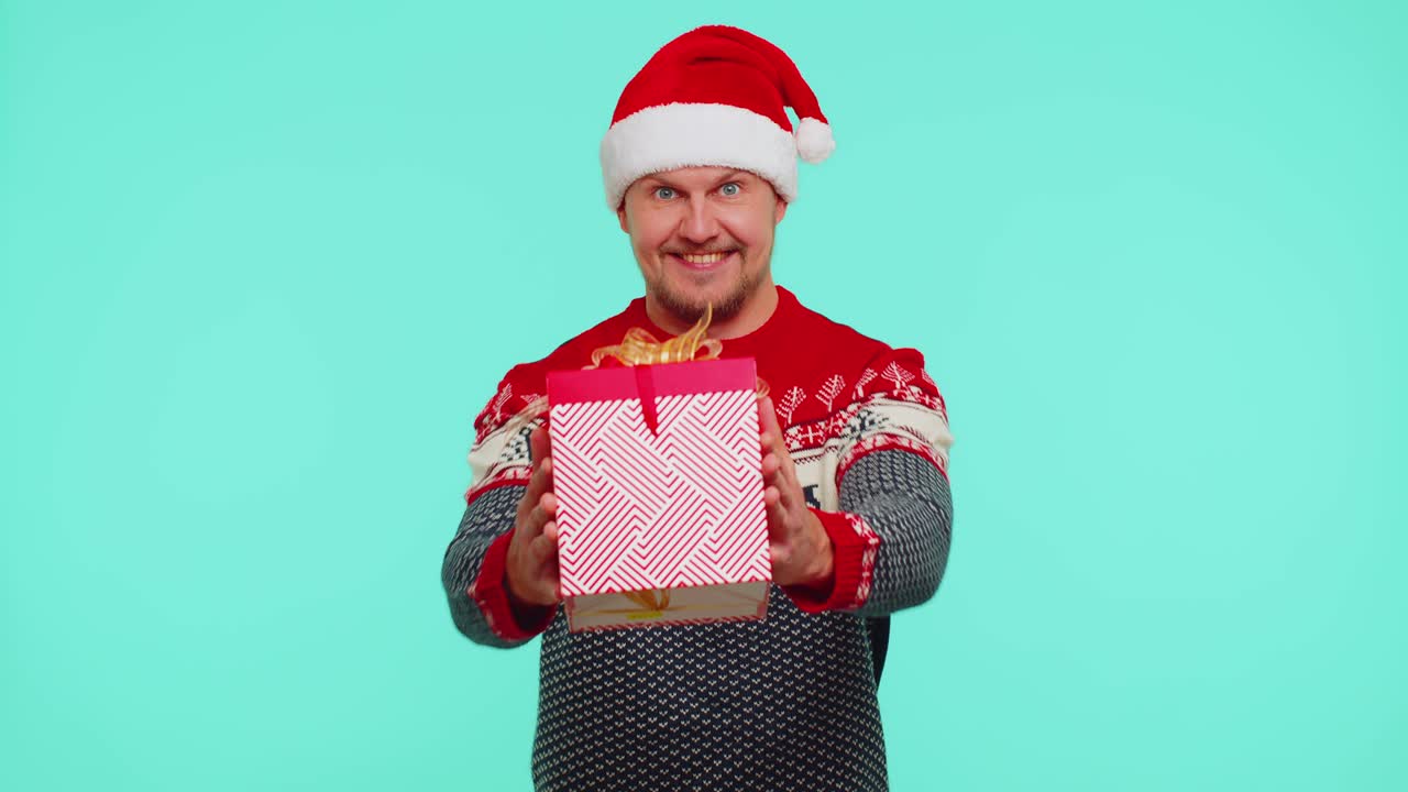 산타 스웨터를 입은 유쾌한 남자가 크리스마스 선물 상자를 선물하고, 카메라로 손을 는다.