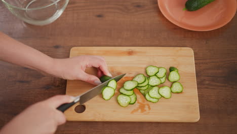 overhead view of person using knife to slice cucumber into thin pieces on wooden cutting board with pile of cucumber slices and plate holding whole cucumber on wooden kitchen counter