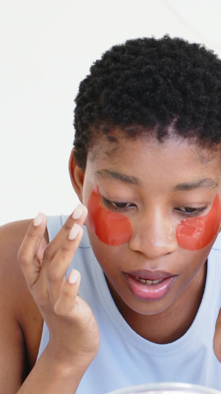 Vertical video: Raising patch, woman placing red gel eye patches at home for skincare using mirror