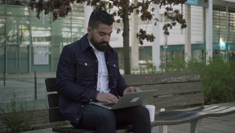 hombre bebiendo de una taza de papel y usando una computadora portátil en la calle