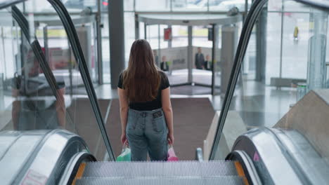 rear view of lady with shopping bags descending on escalator in modern mall, spring water on stone in foreground, clear glass exterior with cars visible outside