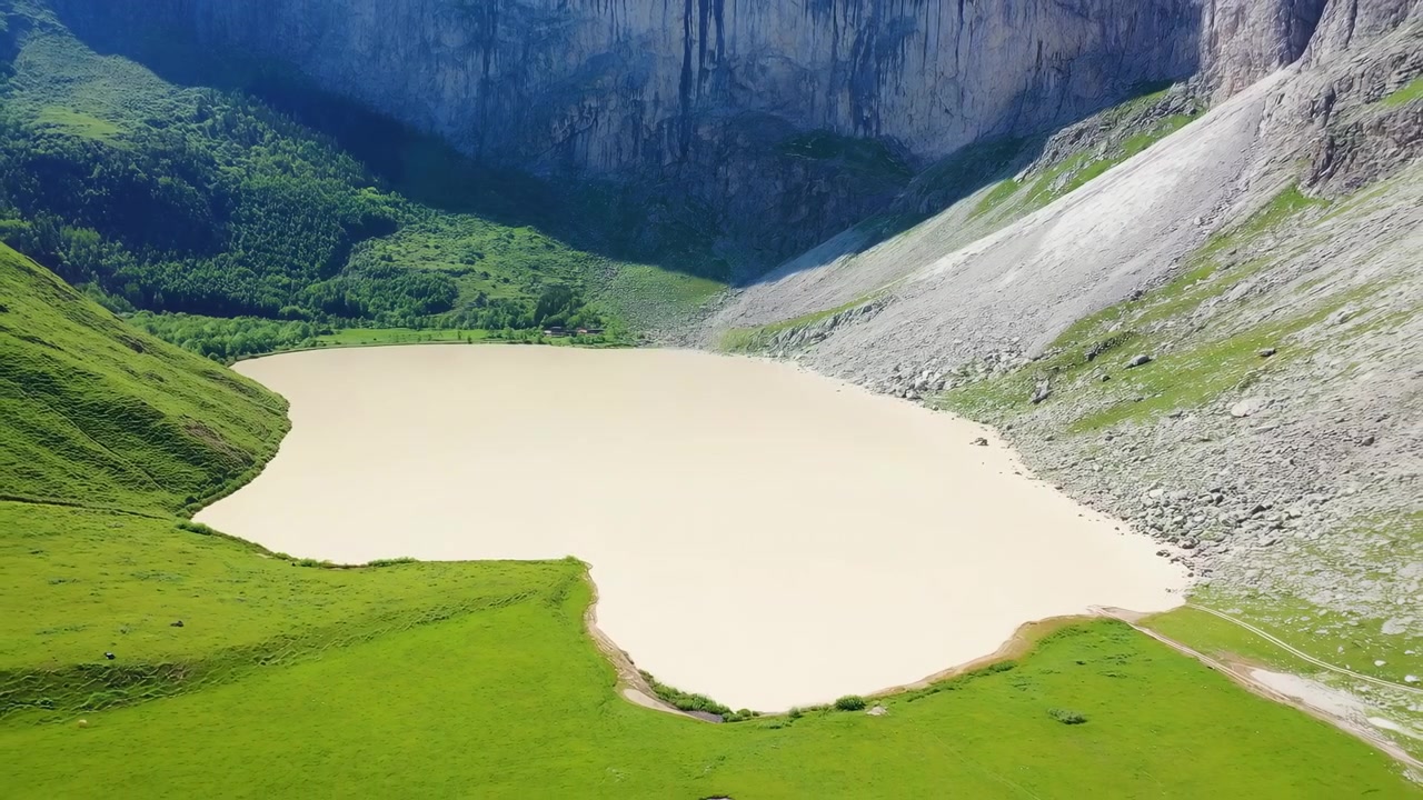 Una veduta aerea a movimento lento rivela un lago craterico pallido circondato da prati vivaci, fitta foresta, pareti rocciose a picco e dolci colline sotto la chiara e luminosa luce del giorno su un terreno montuoso remoto