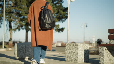 a woman wearing a brown coat, blue jeans, and white shoes walks alone in an urban setting, carrying a black backpack. her posture and slow pace suggest sadness or contemplation