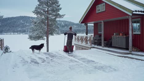 man shoveling snow off in a wood patio of a rustic cottage with his dog in trondheim, norway