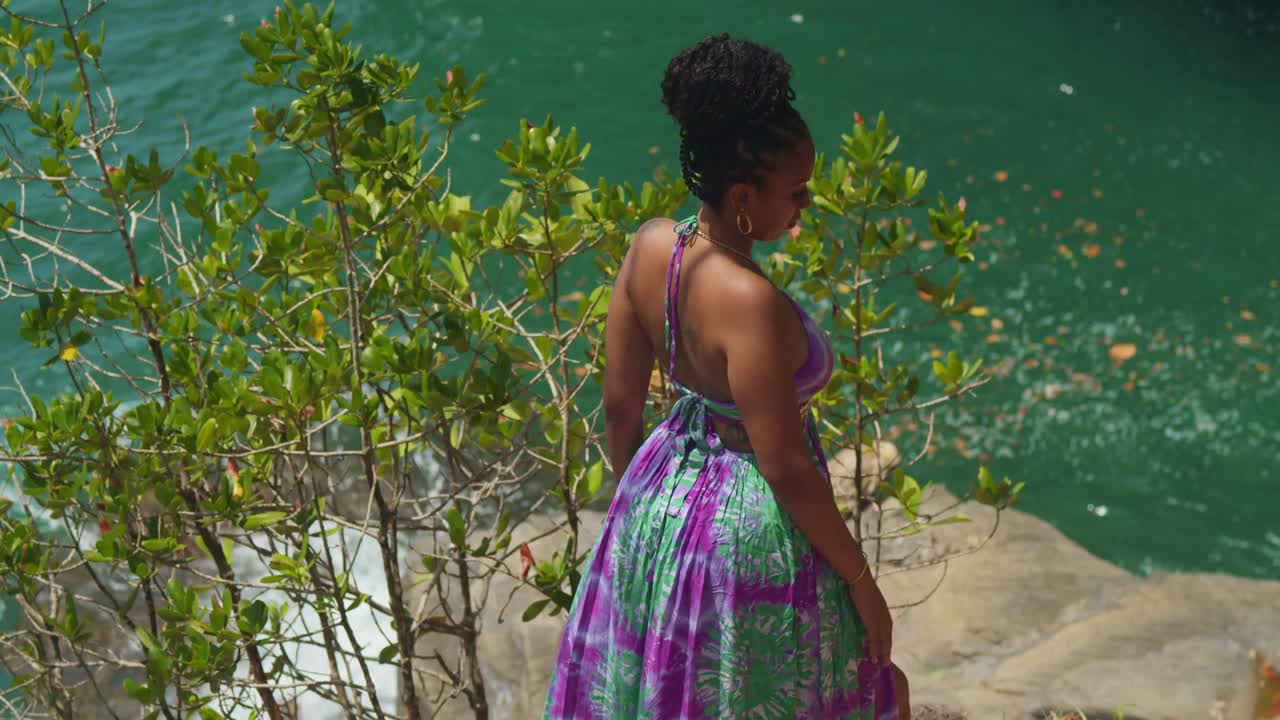 una mujer joven, su largo vestido revoloteando en la brisa, está de pie en un acantilado con vistas a la costa