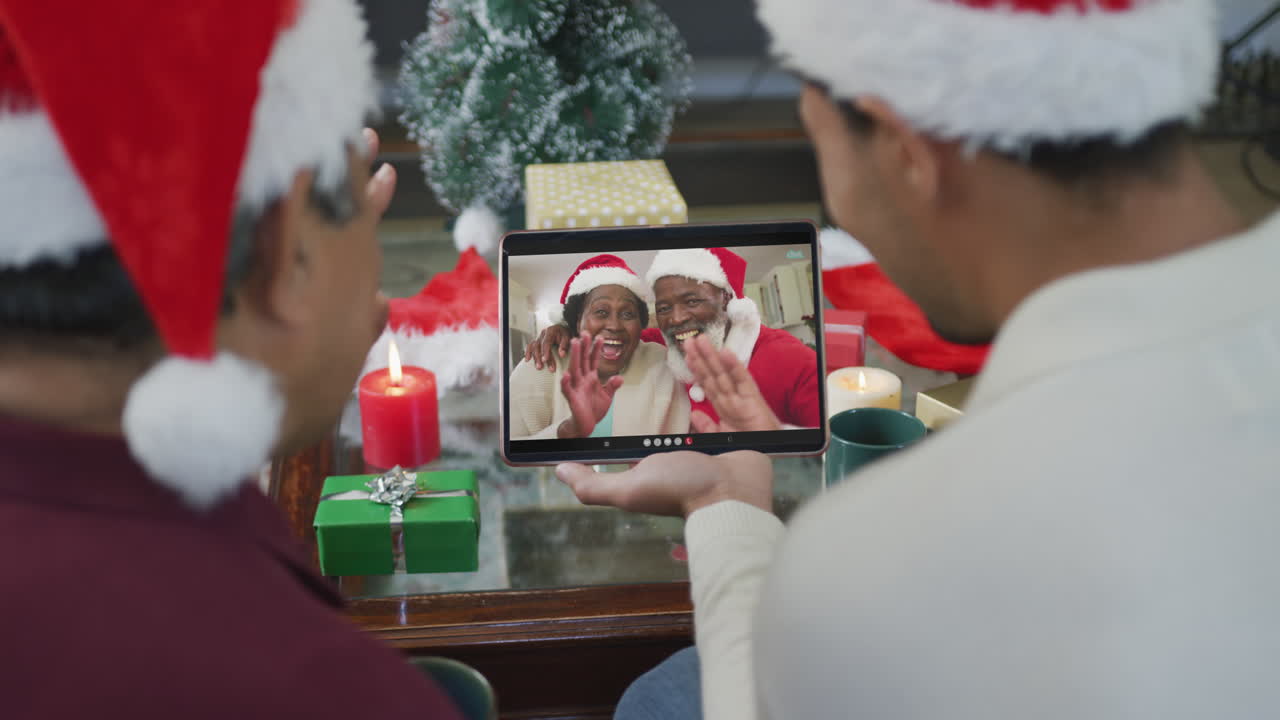 padre y hijo biraciales agitando y usando la tableta para la videollamada de navidad con una pareja feliz en la pantalla