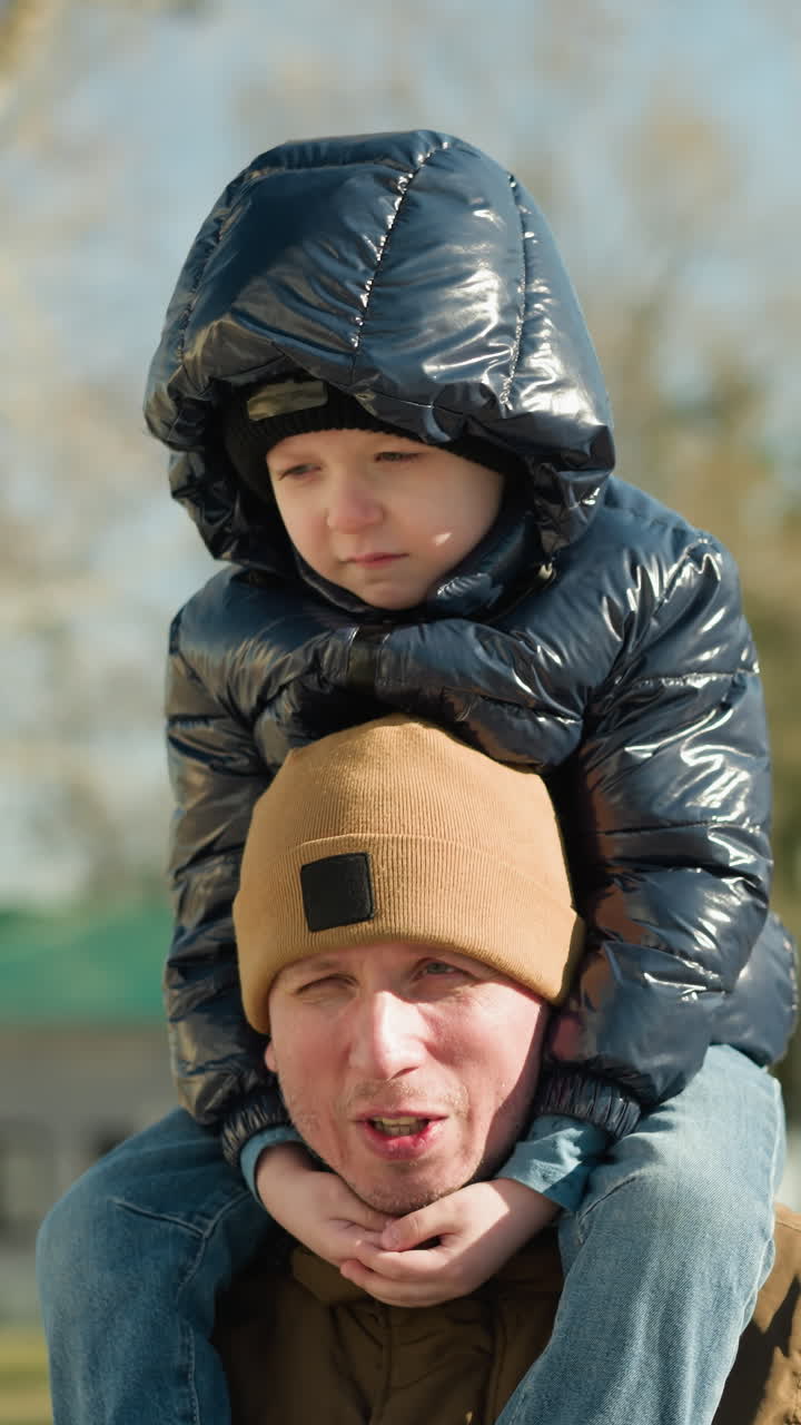 어깨에 아들을 들고 웃고 대화하는 아버지의 클로즈업, 반이는 검은색 재과 후드를 입은 아이, 흐릿한 나무와 빨간색 폴로 셔츠를 입은 사람