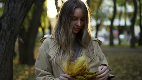 cheerful woman with pile of golden leaves in hands in sunny autumn park