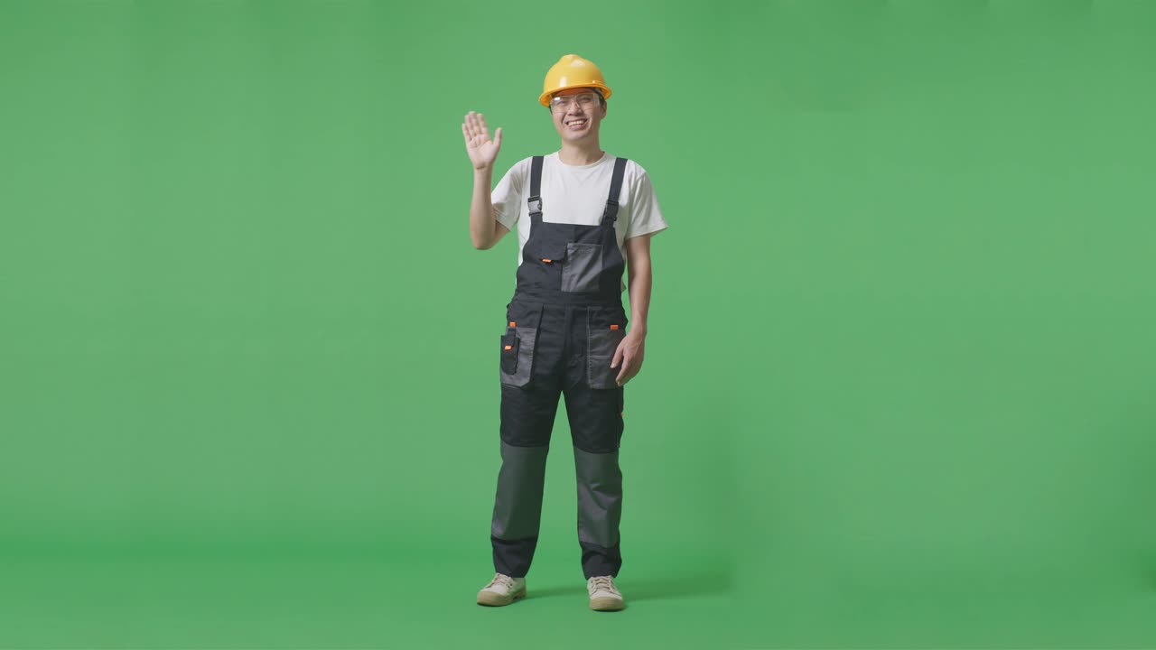 cuerpo lleno de hombre asiático trabajador con gafas de protección y casco de seguridad sonriendo, agitando la mano, y diciendo adiós mientras estaba de pie en el estudio de fondo de pantalla verde