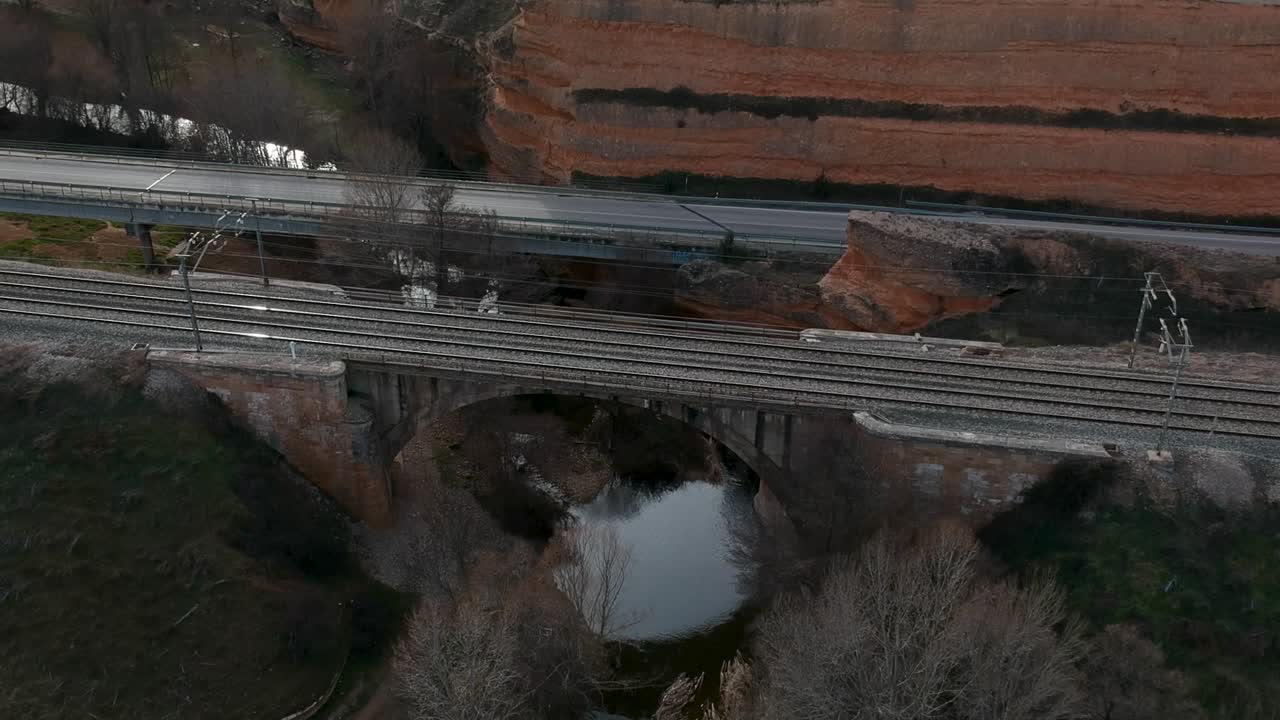 toma aérea moviéndose hacia atrás desde un ferrocarril sobre un río con un tren que pasa a través de él, cerca de un camino de tráfico y un camino de tierra