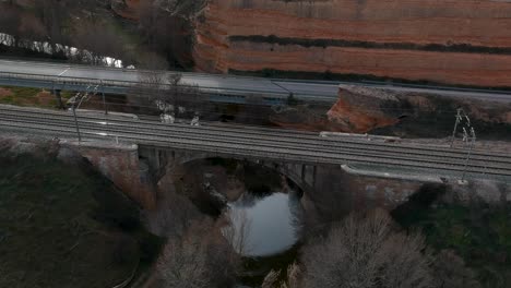 Toma-Aérea-Moviéndose-Hacia-Atrás-Desde-Un-Ferrocarril-Sobre-Un-Río-Con-Un-Tren-Que-Pasa-A-Través-De-él,-Cerca-De-Un-Camino-De-Tráfico-Y-Un-Camino-De-Tierra