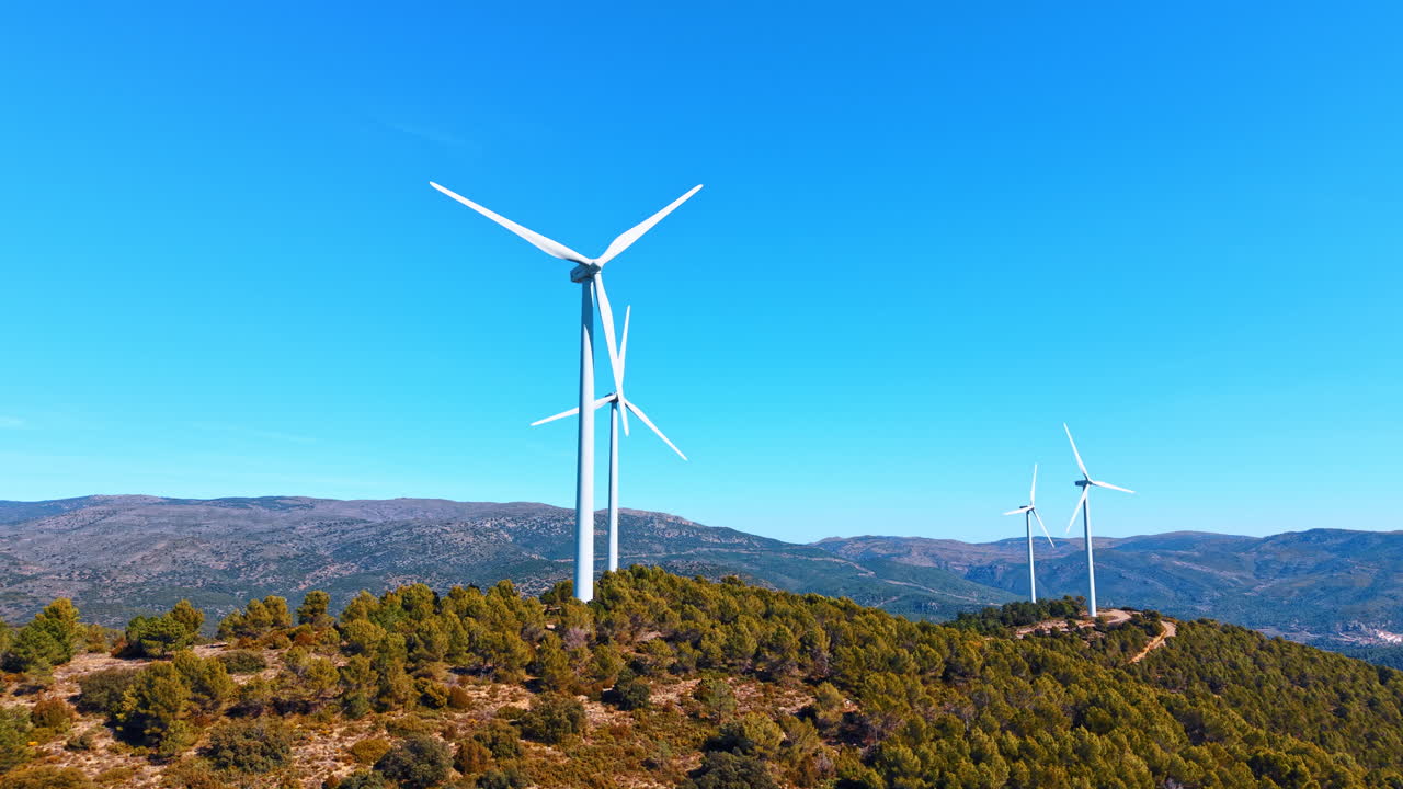 Madri, Espanha, 8 de agosto de 2025: Duas turbinas eólicas em pé no cume de colinas arborizadas e verdes. Perspectiva aérea de geradores eólicos brancos, terreno montanhoso, árvores densas, céu azul, conceito de energia renovável.